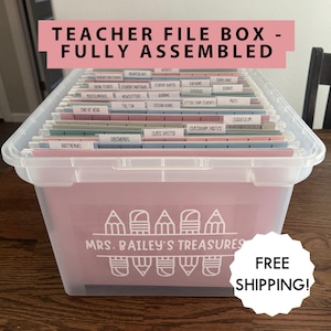 May include: A clear plastic teacher file box with a pink front panel. The box is filled with labeled file folders. Text on the box reads "MRS. BAILEY'S TREASURES." The top of the box has the text "TEACHER FILE BOX - FULLY ASSEMBLED."