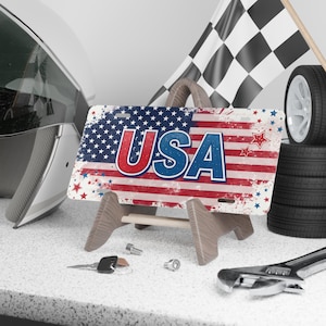 May include: A USA-themed license plate with a distressed American flag design, displayed on a wooden stand. The plate features the word "USA" in bold red and blue letters. A racing flag, tires, and tools are in the background.