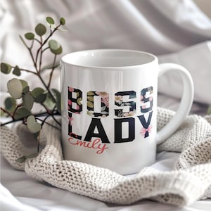 Puede incluir: Taza de cerámica blanca con las palabras "BOSS LADY" en letras negras, con patrones florales en las letras "BOSS". El nombre "Emily" está escrito debajo en cursiva. Un lazo rosa adorna la letra "Y".