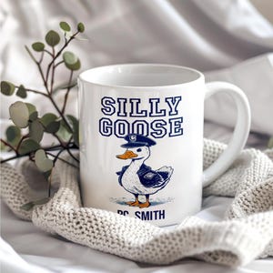 May include: White ceramic mug with the words "SILLY GOOSE" in navy blue lettering. Below the text is a cartoon goose wearing a navy blue hat and jacket. The name "DC SMITH" is printed below the goose.