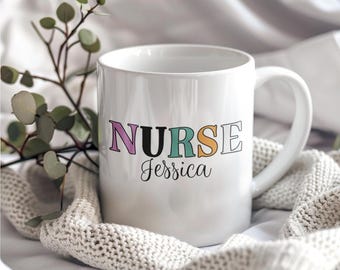 May include: White ceramic mug with the word "NURSE" in colorful letters and the name "Jessica" written below. The mug is sitting on a white and beige knitted blanket.