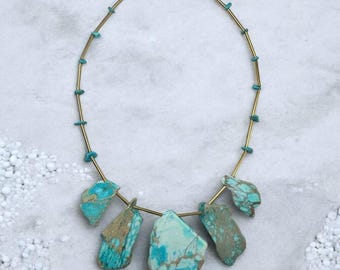 May include: A turquoise and brass necklace with a pendant made of five turquoise stones. The stones are irregular in shape and have a marbled pattern.