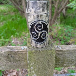 Può includere: Un contenitore cilindrico in legno con una finitura chiara e usurata, caratterizzato da un simbolo triskelion intagliato in scuro sulla parte anteriore. Il contenitore presenta un coperchio abbinato con una piccola linguetta in pelle nera.