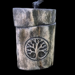 May include: A cylindrical wooden box with a lid, featuring a carved tree of life design within a circular frame. The box has a dark gray and brown wood grain pattern and a leather pull on the lid.