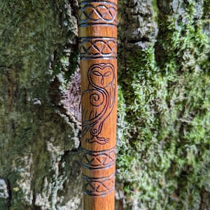 Könnte beinhalten: Ein polierter Gehstock aus Holz mit einem geschnitzten Eulenmotiv auf dem Schaft. Die Eule wird von zwei Bändern mit komplexen, sich wiederholenden keltischen Knotenmustern flankiert, die in das Holz eingraviert sind und eine texturierte Oberfläche bilden.