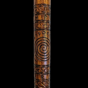 Op de afbeelding: Een gesneden houten wandelstok met gedetailleerde patronen. De stok heeft geometrische banden, een spiraalmotief en mogelijke runen. Het hout is warmbruin, met donkere gravures die een contrast vormen. Ongeveer 150 cm lang.