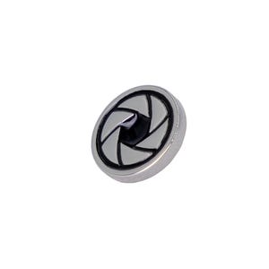 May include: A small, silver-colored button with a black and white aperture design. The aperture symbol is centered on the button, with six curved blades creating the opening. The button has a smooth, metallic finish.