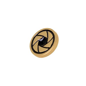 May include: A gold-colored, circular button with a black aperture design in the center. The button has a glossy finish and a raised edge. The aperture design is a series of curved black lines that create a star-like shape.