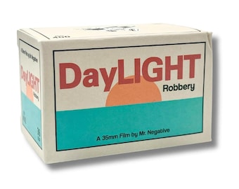 Puede incluir: Una caja rectangular de película de 35mm de Mr. Negative titulada DayLIGHT Robbery. El empaque es blanco con una banda horizontal verde azulado, un gráfico de semicírculo naranja grande y texto en rojo y negro. Indica 400 y 36 exposiciones.