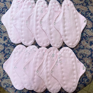 May include: A collection of ten pink, reusable cloth menstrual pads. Each pad features a unique embroidered floral design in shades of pink and green. The pads are oval-shaped with snap closures.