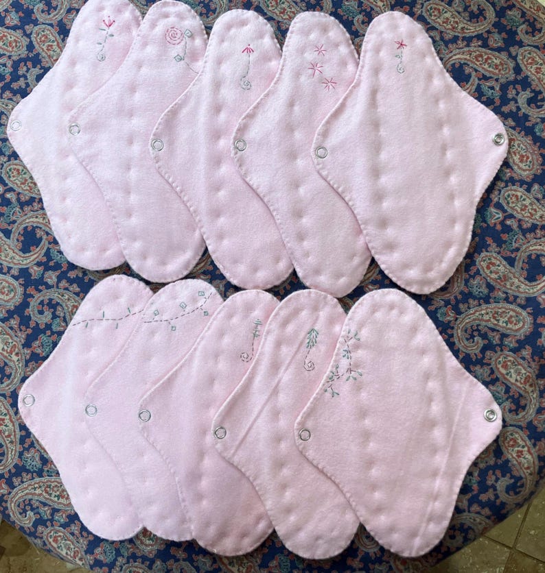 May include: A collection of ten pink, reusable cloth menstrual pads. Each pad features a unique embroidered floral design in shades of pink and green. The pads are oval-shaped with snap closures.