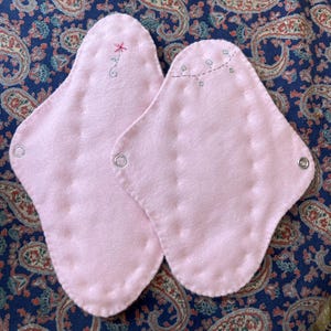 May include: Two pink reusable menstrual pads with decorative stitching and metal snaps. One pad has a small embroidered flower, the other has a line of embroidered squares. The pads are on a blue paisley patterned fabric.