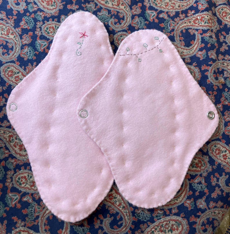 May include: Two pink reusable menstrual pads with decorative stitching and metal snaps. One pad has a small embroidered flower, the other has a line of embroidered squares. The pads are on a blue paisley patterned fabric.
