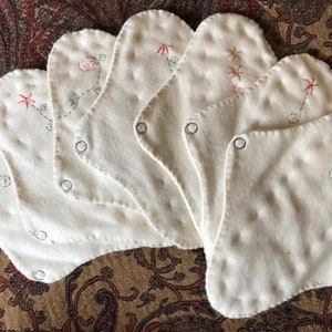 May include: A collection of seven white, petal-shaped cloth pads. Each pad features delicate embroidery of flowers and stars in red and green thread. Metal snaps are visible on each pad.