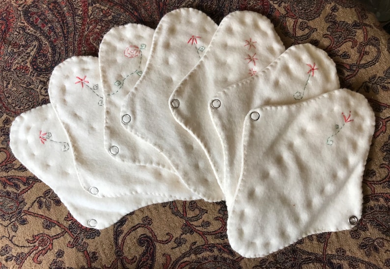 May include: A collection of seven white, petal-shaped cloth pads. Each pad features delicate embroidery of flowers and stars in red and green thread. Metal snaps are visible on each pad.