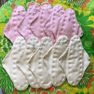 May include: A collection of reusable cloth menstrual pads in shades of pink and cream. Each pad features decorative embroidery and snap closures. The pads are arranged on a floral patterned fabric.