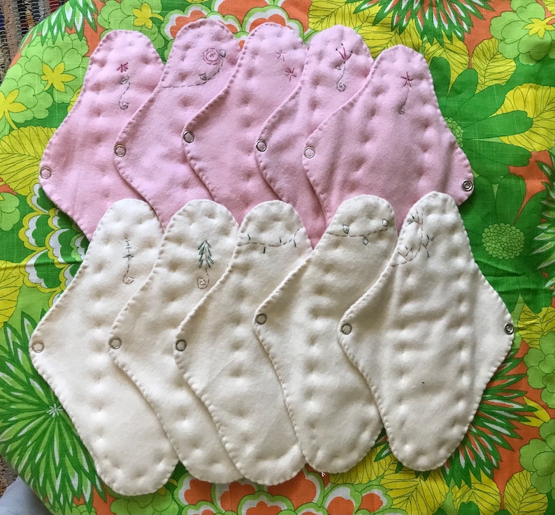 May include: A collection of reusable cloth menstrual pads in shades of pink and cream. Each pad features decorative embroidery and snap closures. The pads are arranged on a floral patterned fabric.