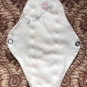 May include: White, reusable cloth menstrual pad with a stitched red rose and green vine design. The pad has a quilted texture and metal snaps on each end for secure fastening. The pad is on a patterned brown background.