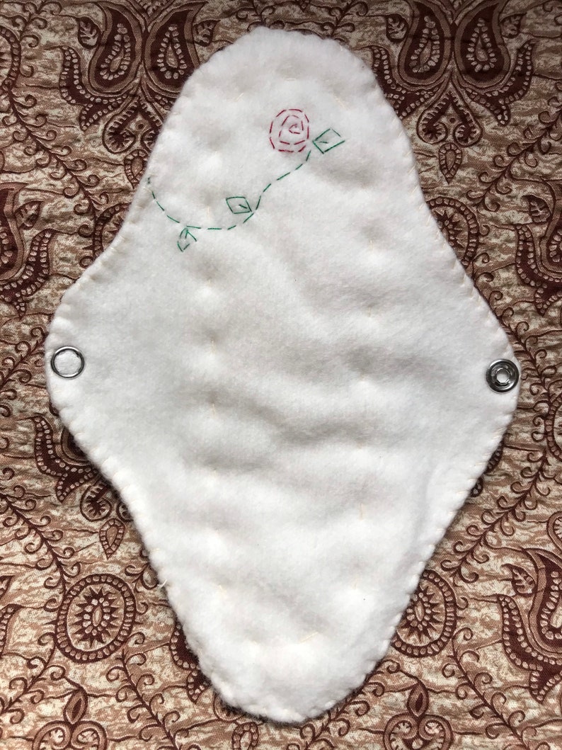 May include: White, reusable cloth menstrual pad with a stitched red rose and green vine design. The pad has a quilted texture and metal snaps on each end for secure fastening. The pad is on a patterned brown background.
