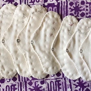 May include: A collection of ten white, reusable cloth menstrual pads with decorative embroidery. Each pad features a different embroidered design in shades of gray and red. The pads are arranged on a purple patterned surface.