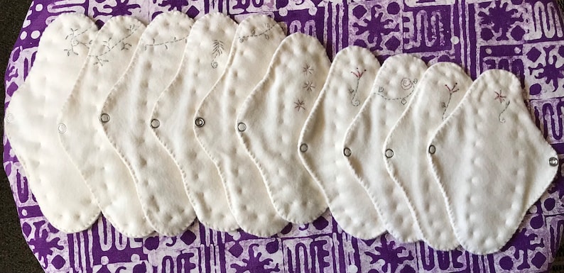 May include: A collection of ten white, reusable cloth menstrual pads with decorative embroidery. Each pad features a different embroidered design in shades of gray and red. The pads are arranged on a purple patterned surface.