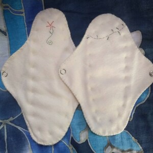 May include: Two off-white, reusable cloth menstrual pads. One pad features a red embroidered flower design, the other has a green vine. Each pad has metal snaps for secure fastening. The pads are displayed on a blue and white patterned fabric.