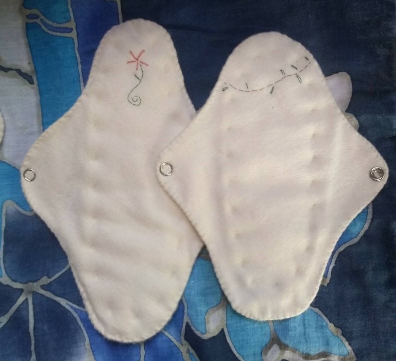 May include: Two off-white, reusable cloth menstrual pads. One pad features a red embroidered flower design, the other has a green vine. Each pad has metal snaps for secure fastening. The pads are displayed on a blue and white patterned fabric.