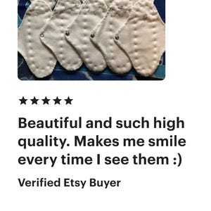 May include: A collection of white, petal-shaped cloth pads with stitched details, arranged on a patterned blue background. The pads are likely reusable menstrual products, designed for comfort and eco-friendliness.