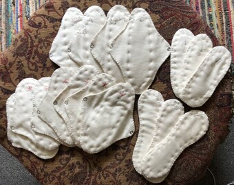 May include: A collection of white cloth menstrual pads with embroidered designs. The pads are arranged in a pile on a patterned fabric surface.