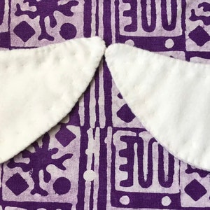 Puede incluir: Dos cuellos de tela blanca con borde festoneado, sobre una tela estampada de color morado y blanco.