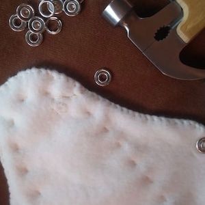 May include: A close-up shot of craft supplies on a brown surface. The image features silver metal eyelets, a hammer, and a white, quilted fabric. The eyelets are scattered, and some are already attached to the fabric.
