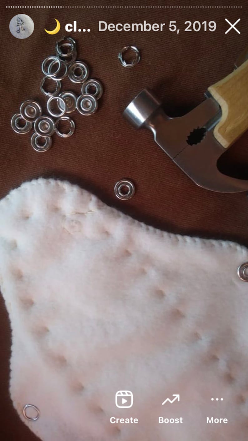 May include: A close-up shot of craft supplies on a brown surface. The image features silver metal eyelets, a hammer, and a white, quilted fabric. The eyelets are scattered, and some are already attached to the fabric.