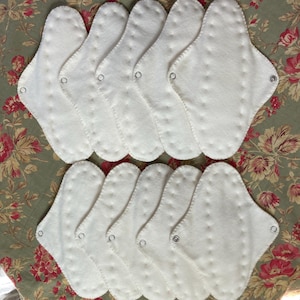 May include: A collection of white, reusable cloth menstrual pads. Each pad is oval-shaped with a scalloped edge and a snap closure. The pads are arranged on a floral patterned fabric.