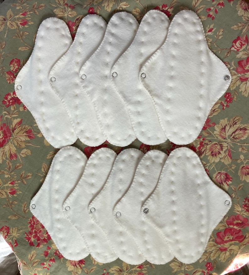 May include: A collection of white, reusable cloth menstrual pads. Each pad is oval-shaped with a scalloped edge and a snap closure. The pads are arranged on a floral patterned fabric.