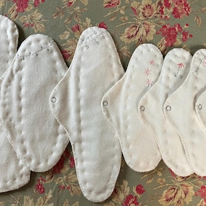 May include: A collection of white, reusable cloth menstrual pads. Some pads feature embroidered floral designs in pink and gray thread. Each pad has a snap closure. The pads are arranged on a floral patterned fabric.