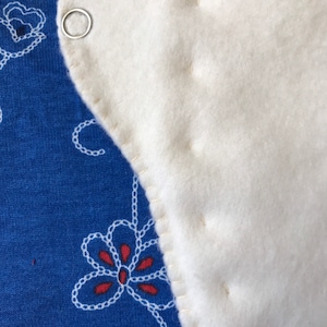 May include: Close-up of a white, rounded fabric piece with a metal ring attached, stitched to a blue fabric. The blue fabric has a floral pattern in white and red.