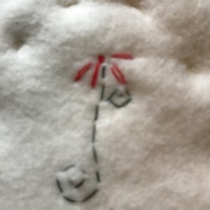 May include: Close-up of a white, fluffy fabric with a hand-stitched design. The design features a red flower with a black stem and a small, geometric shape at the base. The stitching is done with black thread.
