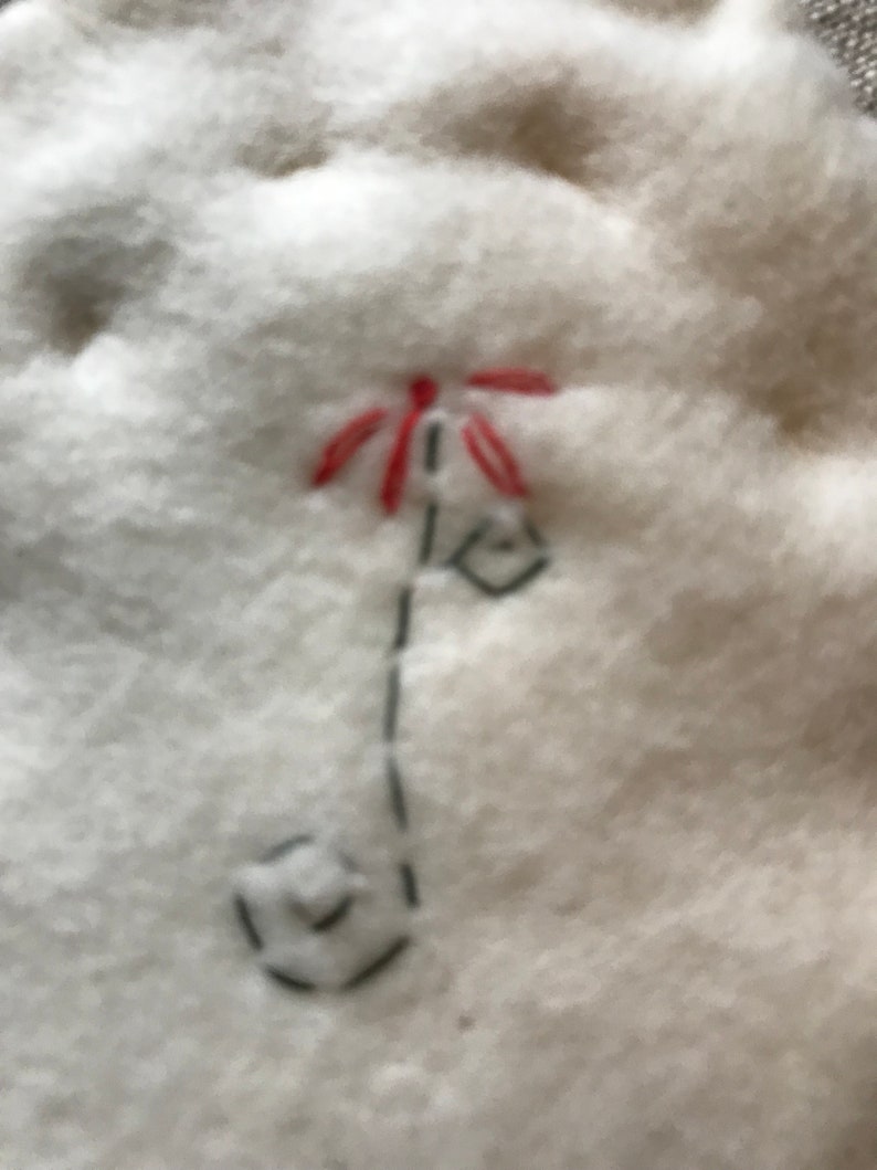 May include: Close-up of a white, fluffy fabric with a hand-stitched design. The design features a red flower with a black stem and a small, geometric shape at the base. The stitching is done with black thread.