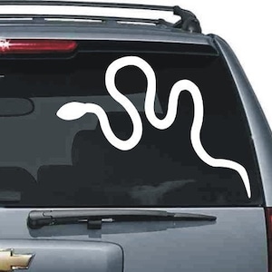 Puede incluir: Calcomanía de serpiente blanca en la ventana de un coche. La calcomanía es un contorno estilizado de una serpiente, con un cuerpo curvado y una cabeza pequeña. La calcomanía está aplicada en la ventana trasera de un vehículo.