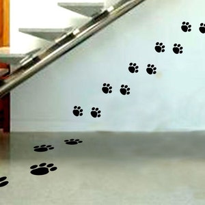Puede incluir: Pegatinas negras con huellas de patas que suben por una escalera y cruzan el suelo. Las huellas están en pares, creando un rastro visual. La escalera tiene escalones blancos y una barandilla metálica.