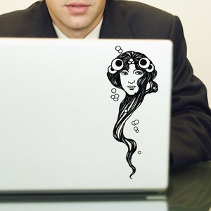 Könnte beinhalten: Ein schwarz-weißes Art-Nouveau-Dekor mit dem Gesicht einer Frau mit langen, fließenden Haaren und dekorativen Kreisen, angebracht auf einem weißen Laptop.