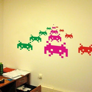 May include: A collection of pixelated alien decals in green, pink, and red are arranged on a light beige wall. The decals are in a retro video game style, creating a playful and nostalgic aesthetic. The wall art adds a pop of color.