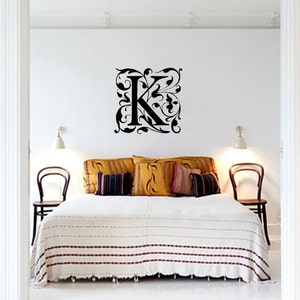 May include: A black, ornate wall decal featuring the letter "K" surrounded by floral patterns. The decal is mounted on a white wall above a bed with yellow and patterned cushions and a white blanket with red stripes. Two chairs are visible.