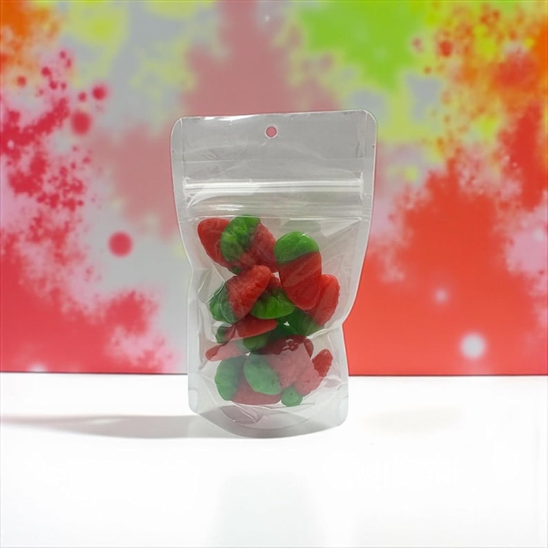 May include: A clear, resealable plastic pouch filled with red and green strawberry-shaped gummy candies. The bag features a zip-lock closure at the top and a small circular hole for hanging, providing a clear view of the colorful fruit-shaped treats.