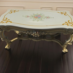 May include: An ornate, antique coffee table with a cream-colored top and gold accents. The tabletop features a floral design in the center and gold embellishments on the edges. The table has intricately carved legs and a decorative apron.