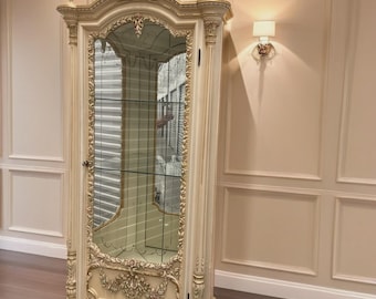 Könnte beinhalten: Ein hoher, cremeweißer Vitrinenschrank mit verzierten goldfarbenen Schnitzereien, einer einzelnen Glastür mit verspiegelter Rückwand und mehreren Glasböden im Inneren. Das Möbelstück ist mit dekorativen Zierknöpfen und floralen Ornamenten versehen.