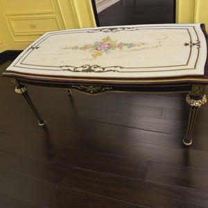 May include: Rectangular coffee table with a cream-colored top featuring a floral design in pink, yellow, and blue. The table has a dark brown border, gold accents, and ornate legs. The table is approximately 48 inches long.