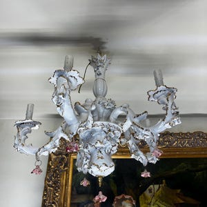 May include: A white porcelain chandelier with gold accents and pink flower details. The chandelier has six arms and is hanging from a ceiling.