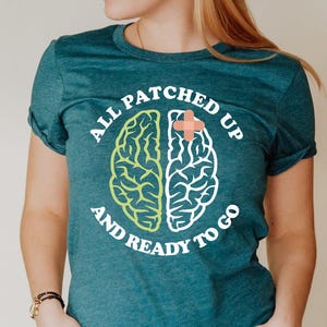 May include: Teal t-shirt with the text "ALL PATCHED UP AND READY TO GO" surrounding a brain graphic. The brain graphic is split in half, with one side green and the other white, and a red cross in the middle.