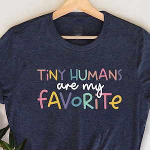 May include: A navy blue t-shirt with the text "TINY HUMANS are my FAVORITE" in colorful letters. The shirt is hanging on a wooden hanger against a white background. The shirt has rolled-up sleeves.